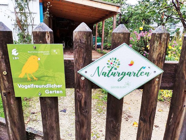 Ein Gartenzaun aus Holz. Daran zwei Schilder. Eines mit dem Titel Naturgarten. Das andere mit der Auszeichnung vogelfreundlicher Garten.
